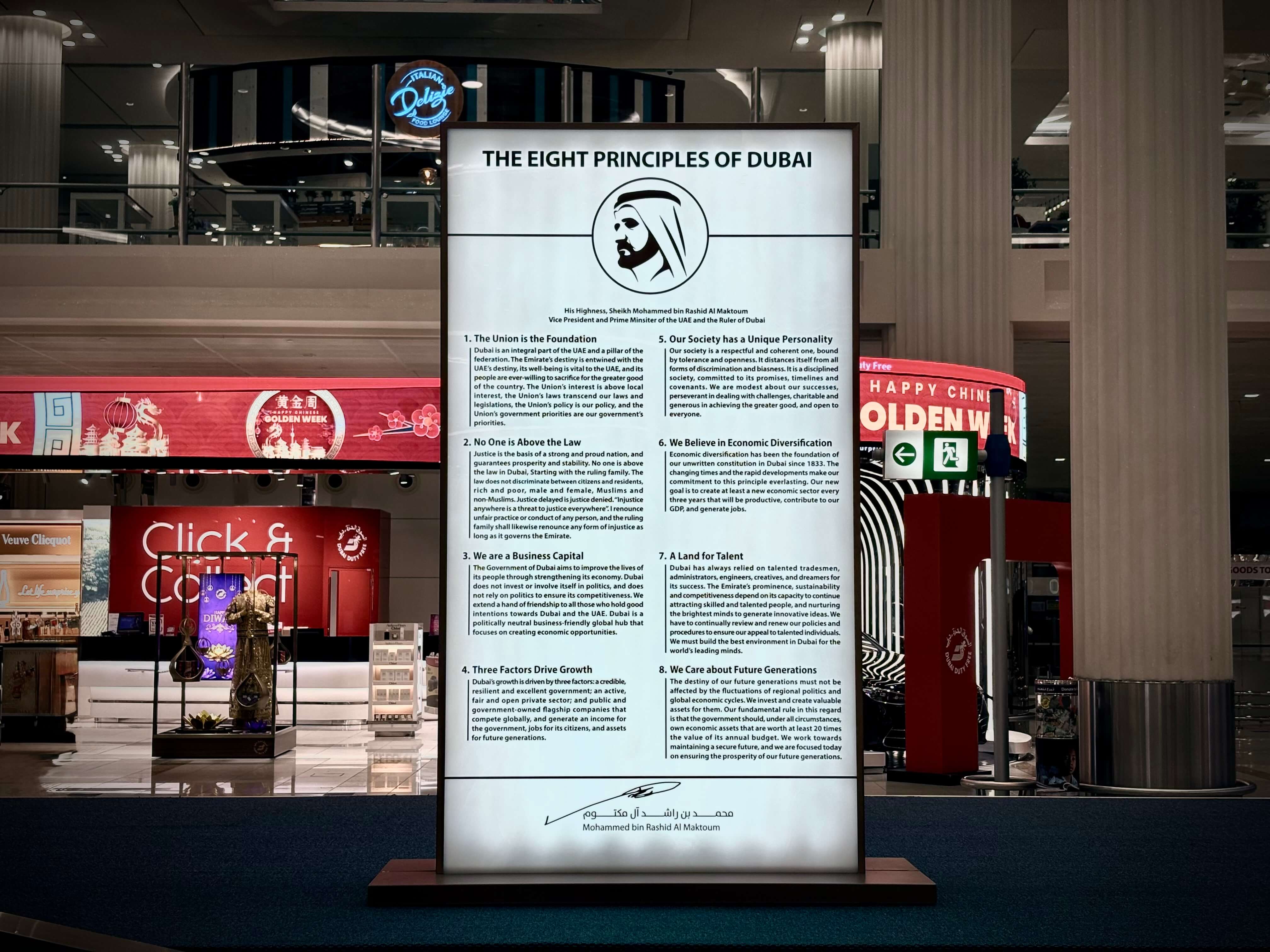 Illuminated sign with ‘The eight principles of Dubai’ at Dubai International Airport, surrounded by shops and pillars