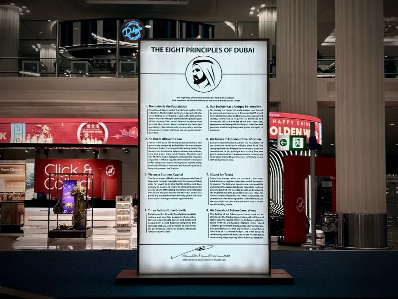 Placa iluminada com Os oito princípios de Dubai no Aeroporto Internacional de Dubai, rodeada por lojas e pilares